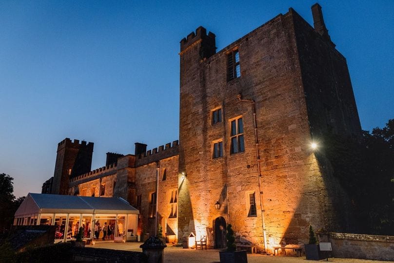 Sudeley Castle in Gloucestershire floodlit at dusk, with a lit marquee alongside the medieval tower